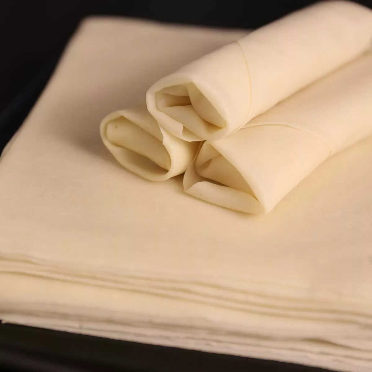 A stack of spring roll sheets with three rolled by hand on top